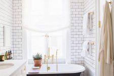 an airy modern bathroom clad with white subway tiles, with a black clawfoot tub, a white vanity with gold touches and gold fixtures