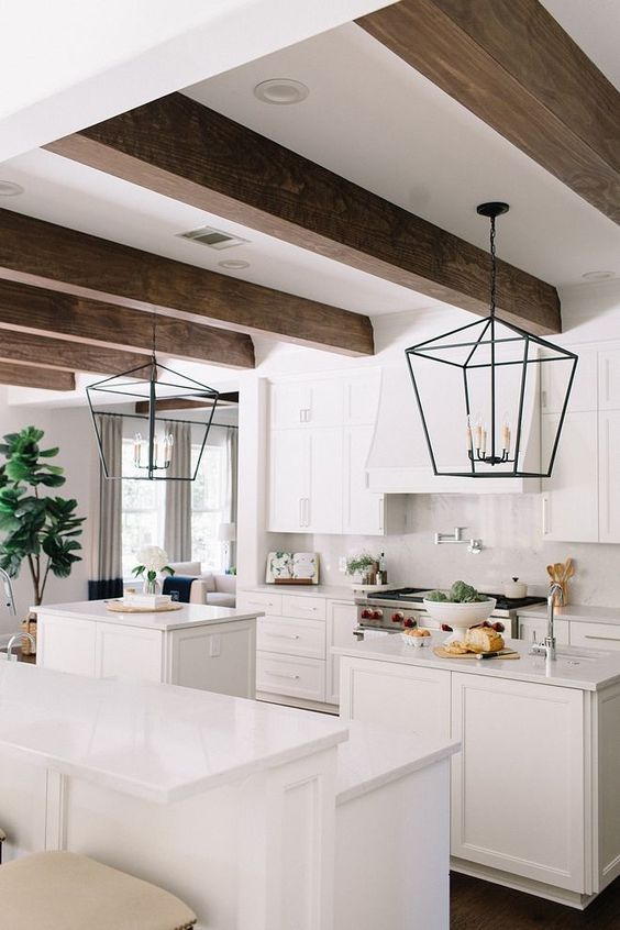 an airy white kitchen with shaker cabinets, a hood, stained wooden beams and two small kitchen islands plus pendant lamps
