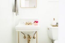 an all-white powder room with a console sink, a brass trash bin, a mirror in a brass frame and a mini gallery wall