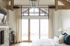 an attic cabin bedroom with wooden beams on the ceiling, a stone clad fireplace, a neutral bed with neutral bedding and a black dresser