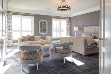 an eye-catchy grey bedroom with an upholstered bed, a creamy sofa, grey chairs, a round table, a chic chandelier and touches of gold