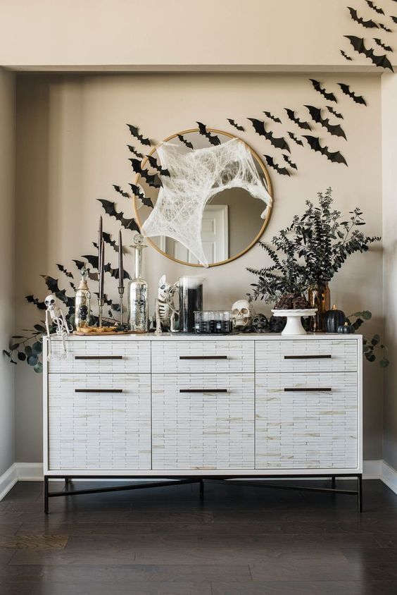 a white console table styled for Halloween, with skeletons, skulls, bones, black paper bats on the wall and a round mirror with spider web