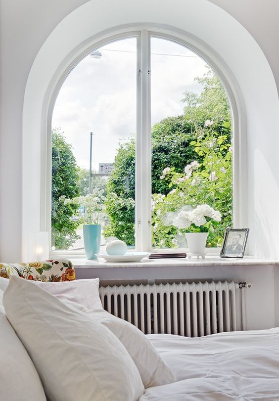 a Scandinavian bedroom with an arched window with a vertical frame, with a windowsill used for potted blooms and an artwork
