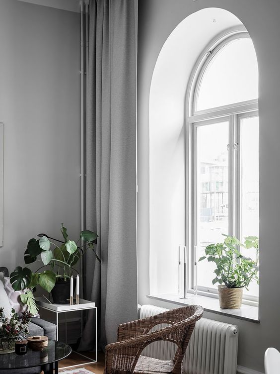 a Scandinavian room with an arched window with a white frame, grey curtains and grey furniture, a woven chair and some greenery