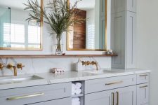 a beautiful dove grey bathroom with shaker style cabinets, white stone countertops and a backsplash, mirrors in brass frames, pendant bulbs, brass fixtures and handles