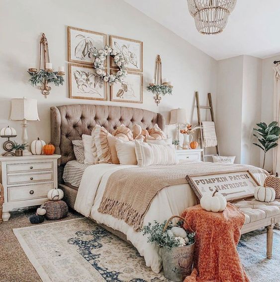 a beautiful farmhouse bedroom with a taupe upholstered bed, matching neutral nightstands, neutral bedding, an upholstered bench, orange touches, faux pumpkins and greenery