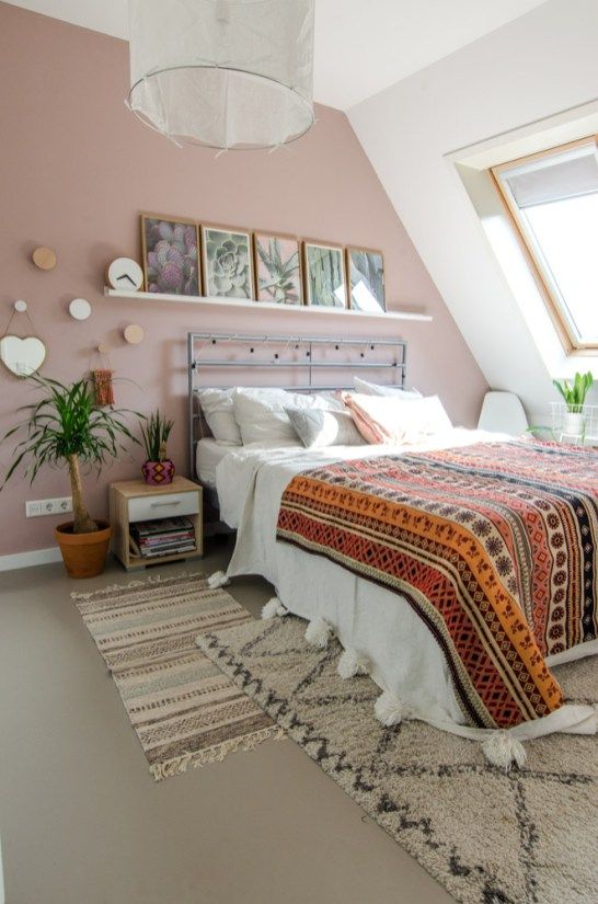 a boho attic bedroom with a dusty pink accent wall, a metal bed with neutral and bright boho bedding, nightstands, potted plants and a gallery wall
