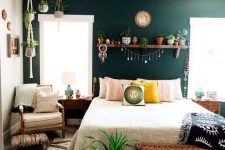 a boho bedroom with a dark green accent wall, a bed, a woven bright bench, a neutral chair, a shelf with plants and more hanging planters