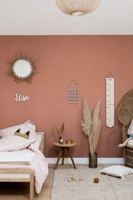a boho kid bedorom with a pink accent wall, a wooden bed, nightstand, a peacock chair, pampas grass, some pretty decor and neutral textiles