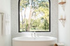 a bold bathroom with marble tiles and an arched window for an accent, an oval tub, a beaded chandelier and a rope side table