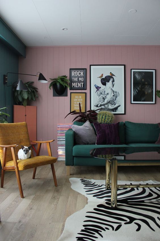 a bold living room with a pink planked wall, a green sofa, a gallery wall, a mustard chair, a zebra rug, potted plants and black sconces