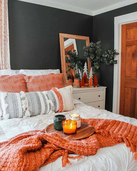 a bright fall bedroom with creamy and white furniture, orange blankets and pillows, a mirror in an orange frame and orange trees and fall scented candles