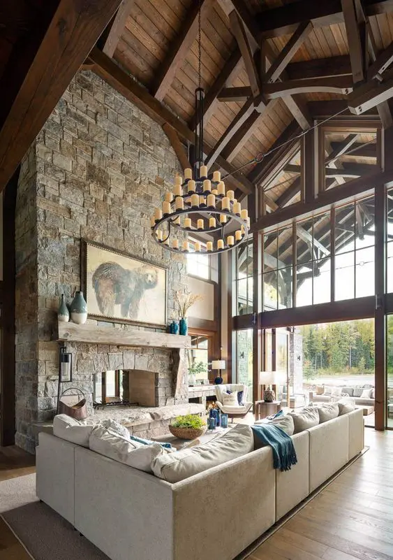 a cabin living room with a tall ceiling with wooden beams, a large fireplace clad with stone, neutral seating furniture and a chandelier