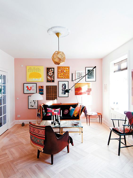 a colorful living room with a pink accent wall, a bold and cool gallery wall on it, an orange sofa with bright pillows, a burgundy chair and a faceted pendant lamp