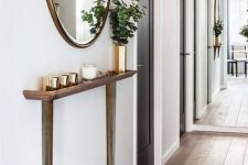 a contemporary entryway with a super sleek console with brass legs, brass jar candles and a vase, a round mirror in a brass frame is cool