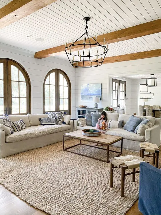 a cool coastal living room with light-stained wooden beams, neutral seating furniture, blue pillows, a coffee table and a large jute rug