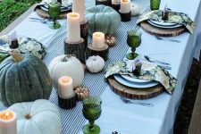 a cool rustic Thanksgiving tablescape with a striped runner, heirloom pumpkins, pillar candles, green glasses, wood slices and floral napkins