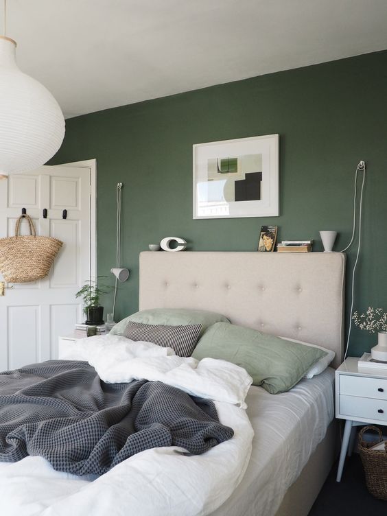 a cozy bedroom with a dark green accent wall, a greige upholstered bed, white nightstands, an artwork and some neutral bedding plus a pendant lamp