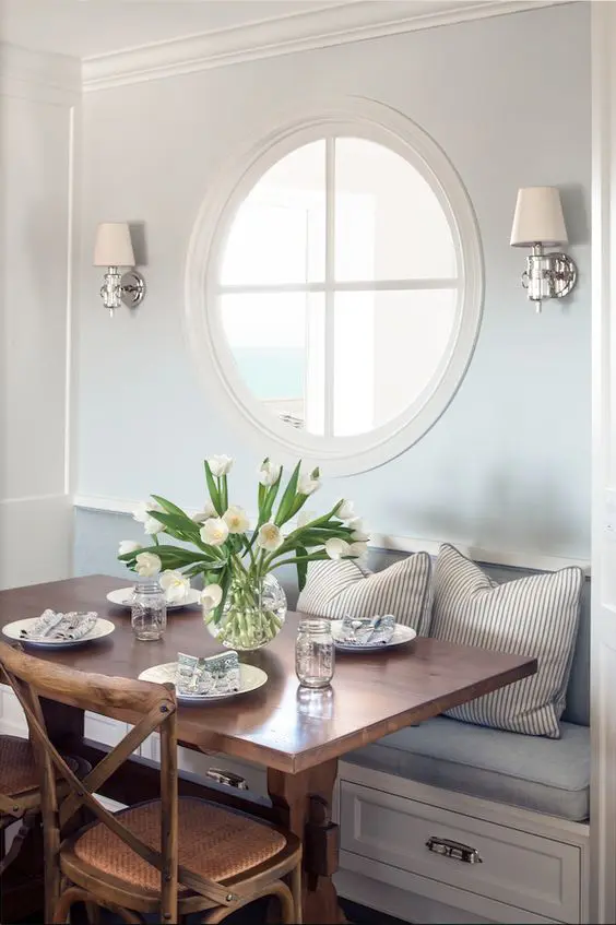 a cozy coastal dining nook with a porthole window and wall lamps looks very refined and very chic