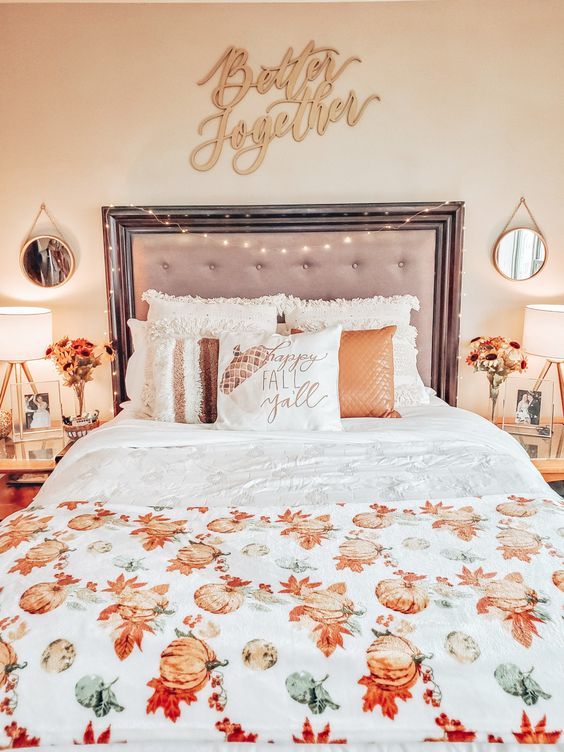 a cozy fall bedroom with an upholstered bed, neutral and orange bedding, a pumpkin blanket, matching nightstands and fall blooms plus elegant table lamps
