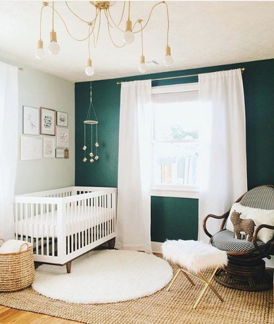 a cozy farmhouse nursery with a dark green accent wall, a white crub and curtains, a dark stained chair, layered rugs and a gilded chandelier