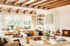 a cozy neutral living room with a fireplace, neutral seating furniture, a large square coffee table and wooden beams, colorful pillows