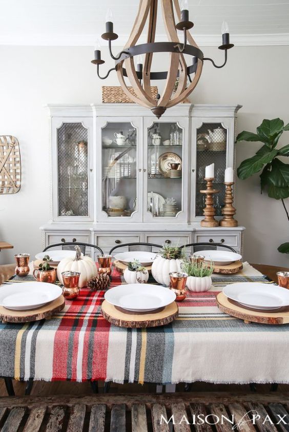 a cozy rustic Thanksgiving table setting with a plaid tablecloth, pumpkins, pinecones, copper mugs, greenery and wood slice placemats