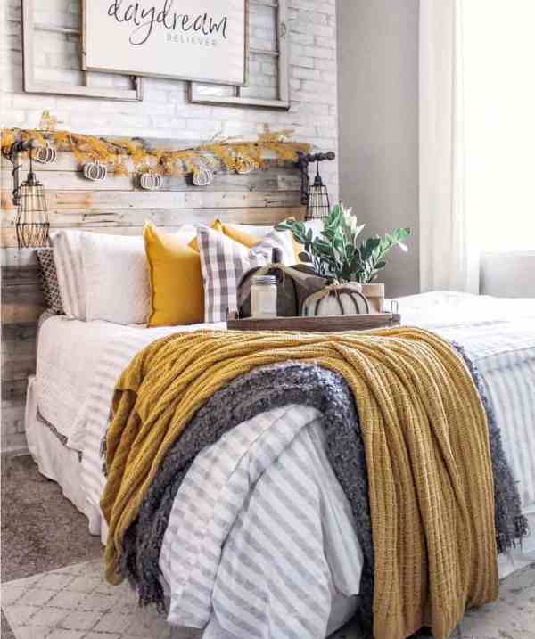 a cozy rustic bedroom with a faux brick accent wall, a reclaimed wooden bed, neutral and mustard bedding, a bright leaf and pumpkin garland and some fabric pumpkins