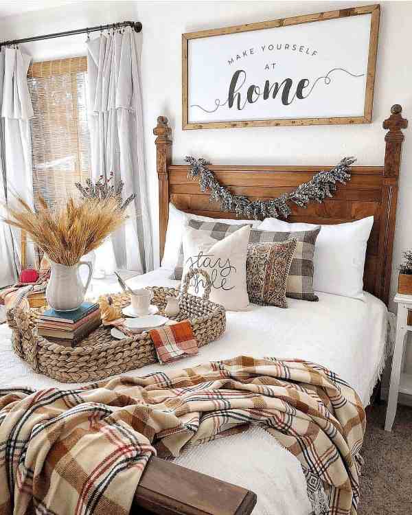 a cozy rustic bedroom with a stained wood bed, neutral and plaid bedding, a fall sign, a basket tray with wheat and vintage books is a very cozy space
