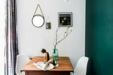a dining space with a dark green accent wall, a stained table, white chairs, a black and white tiled floor and a pendant bulb plus a mini gallery wall