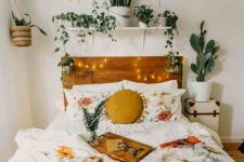 a fall boho bedroom with a stained bed with neutral and printed bedding, a shelf with potted plants, a mustard pillow and lights is a very cozy space