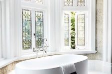 a farmhouse bathroom with a bow window with stained glass, a large oval tub and a printed rug plus lotsof greenery seen through the windows