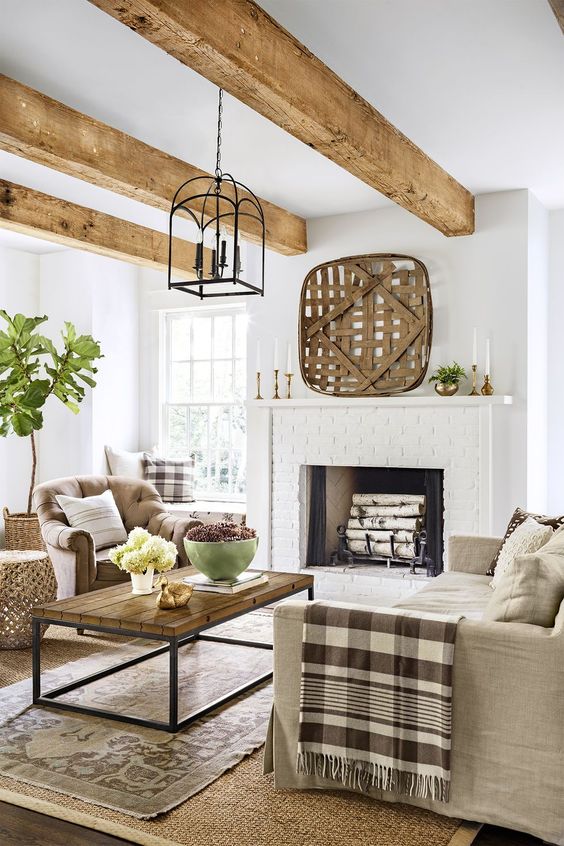 a farmhouse living room with a large brick clad fireplace, neutral seating furniture, a wooden coffee table, wooden beams and layered rugs