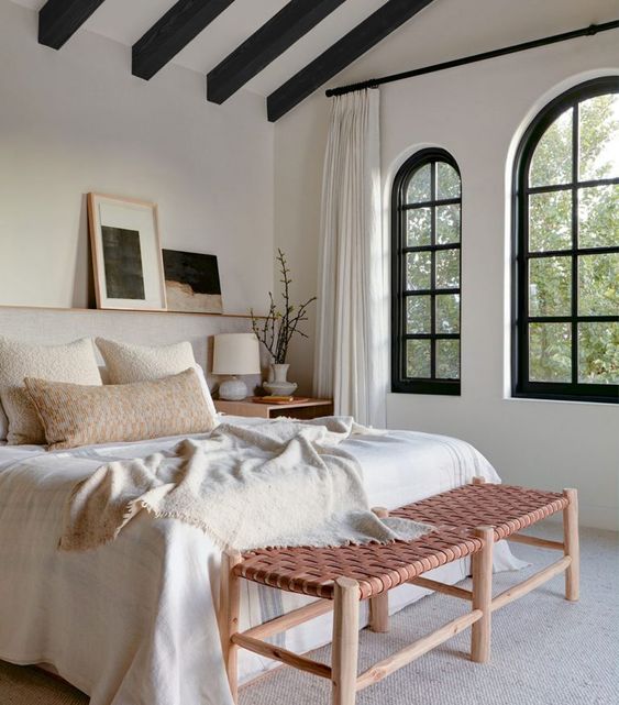 a fresh modern attic bedroom with black framed arched windows and black beams, a neutral bed and a pink woven bench, some art