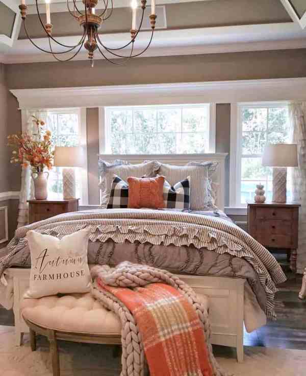 a greige farmhouse bedroom with a white bed, a white upholstered bench, stained nightstands, an orange pillow, an orange plaid blanket and fall leaves
