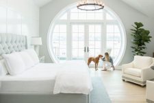 a large modern farmhouse bedroom with paneling, a grey bed with neutral bedding, creamy chairs, a chandelier and a porthole window that includes a door