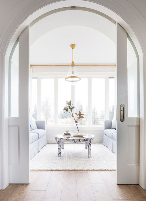 a light-filled white living room with a glazed wall, pale blue sofas and a butterfly print table, white arched pocket doors