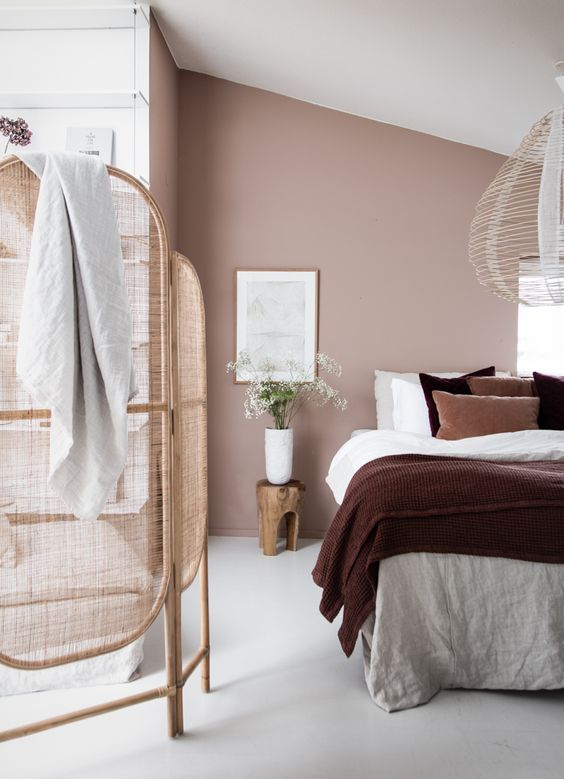 a lovely attic bedroom with a dusty pink accent wall, a neutral bed with bold bedding, a pendant lamp, rattan space divider is very cool