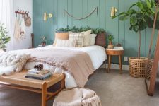 a lovely boho bedroom with a green paneled wall, a plywood bed, a woven bench, round plywood nightstands, potted plants and gilded touches