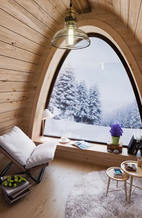a lovely chalet nook with an arched window, everything clad with wooden planks, a comfy chair and some tables