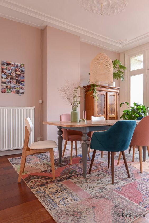a lovely eclectic dining room with a blush accent wall, a vintage dining table, mismatching chairs, potted plants, a woven pendant lamp
