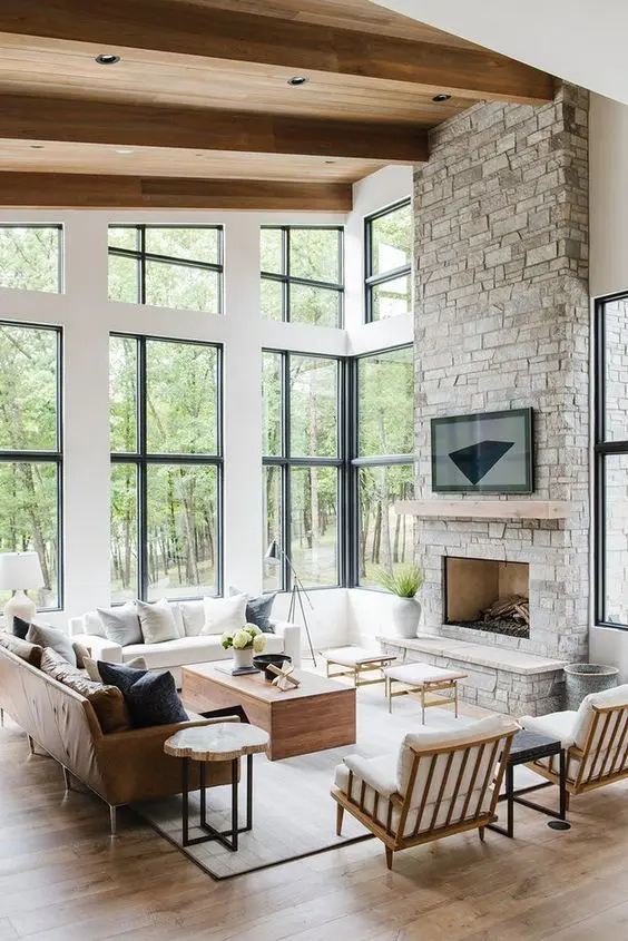 a lovely light filled farmhouse living room with wooden beams, a fireplace clad with stone, neutral seating furniture, coffee tables and double height windows