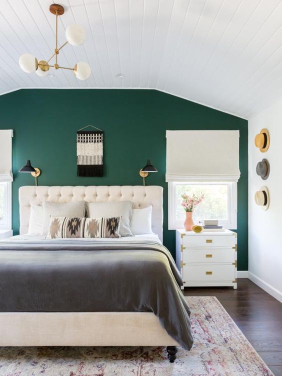 a mid-century modern bedroom with a green accent wall, a creamy upholstered bed, creamy nightstands, a chandelier, some sconces and hats on the wall