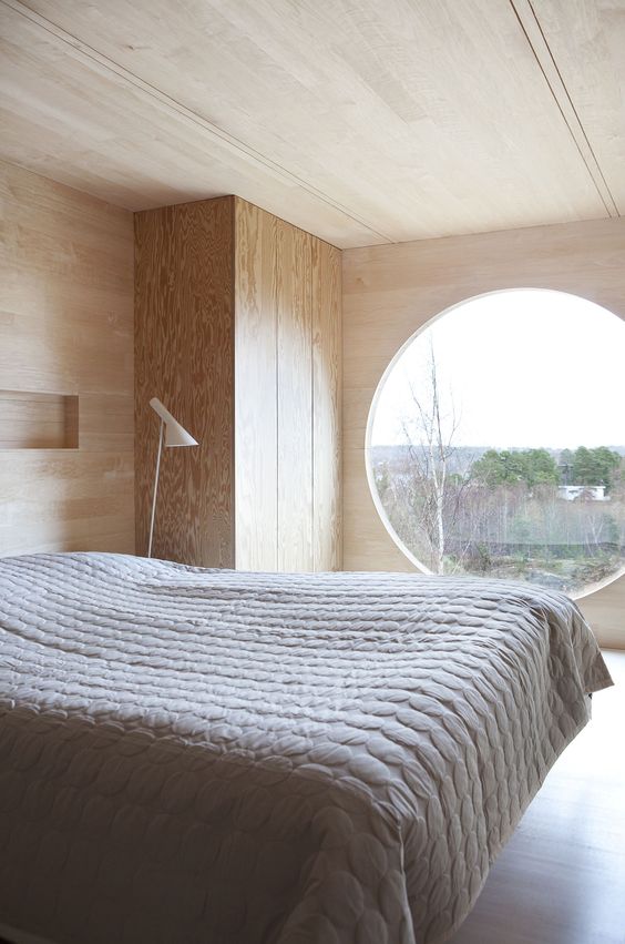 a minimalist bedroom totally clad with plywood, with a large round window, a sleek plywood wardrobe and a bed with neutral textiles