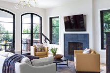 a modern living room with black framed arched windows and doors, a built-in fireplace, a grey sofa and tan chairs plus a bubble chandelier