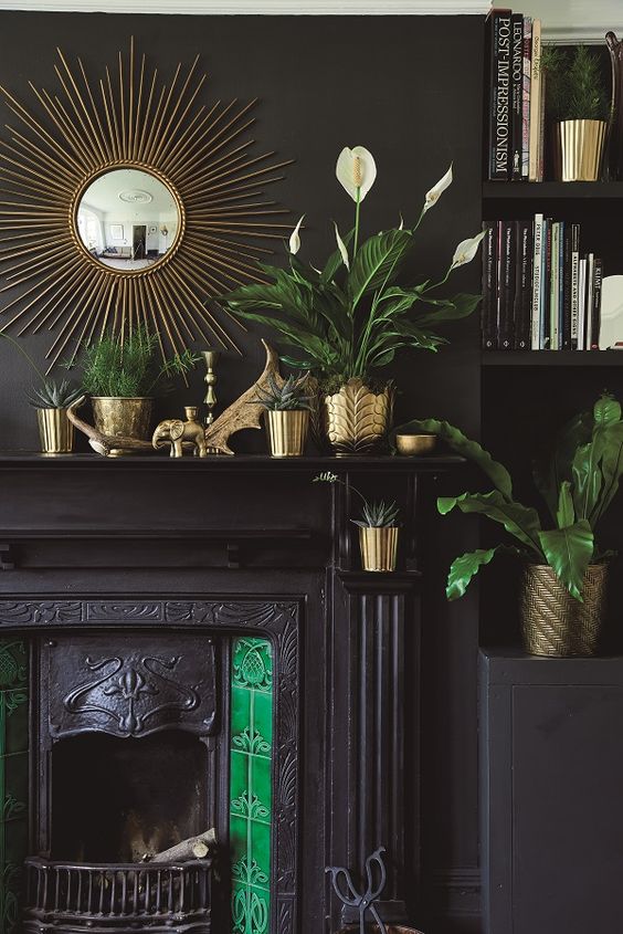 a moody living room with black walls and built in shelves, a built in black fireplace, green tiles, brass planters and pots and greenery in them plus a sunburst brass mirror