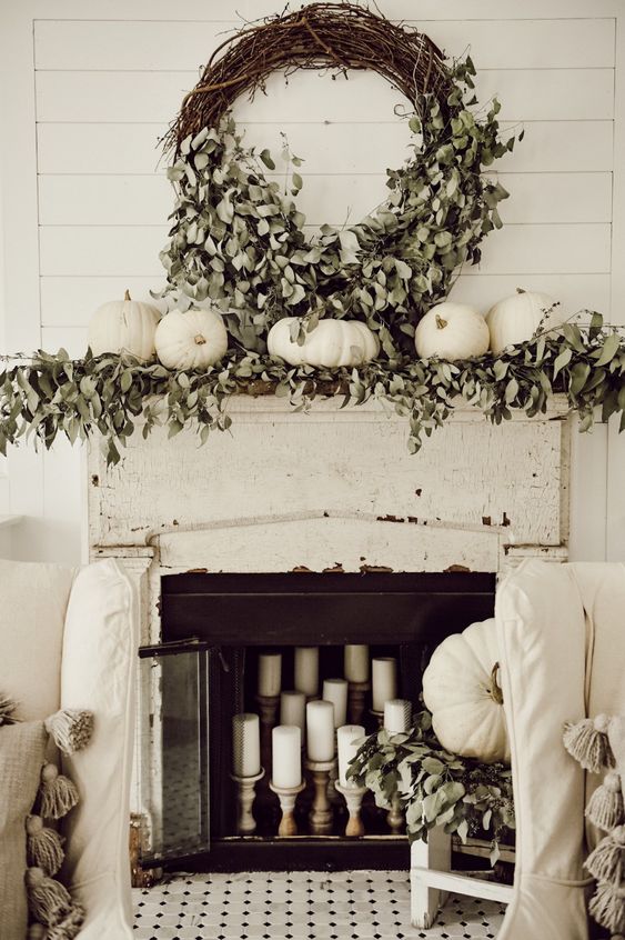 a neutral farmhouse mantel with lush greenery, white pumpkins, a vine wreath with eucalyptus, pillar candles right in the fireplace