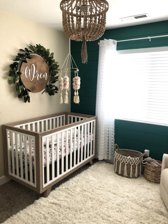 a neutral farmhouse nursery with an emerald planked accent wall, neutral crib, baskets for storage, a wooden bead chandelier and a sign