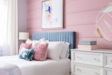 a pastel bedroom with a pink planked wall, a blue bed, white, blue and pink bedding, a white dresser, a mirror in a sheer frame and a sunburst pendant lamp