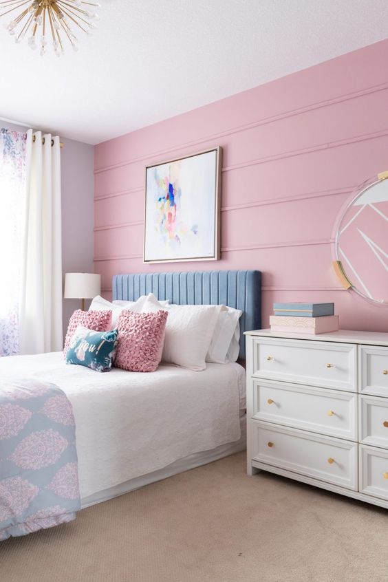 a pastel bedroom with a pink planked wall, a blue bed, white, blue and pink bedding, a white dresser, a mirror in a sheer frame and a sunburst pendant lamp